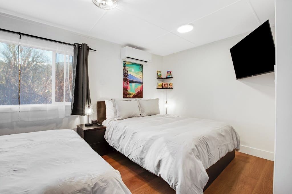 Quarto de hotel com duas camas, uma janela grande com cortinas, uma TV de tela plana presa à parede e um quadro sobre as camas.