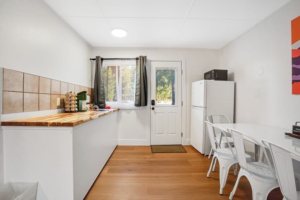 Una cocina compacta con encimera de madera, armarios blancos, un refrigerador y una zona de comedor con sillas blancas.