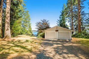 A single-story house with a garage, surrounded by tall trees and a clear sky. - Updated 3BR Bayview - Balcony - Firepit (Allyn)