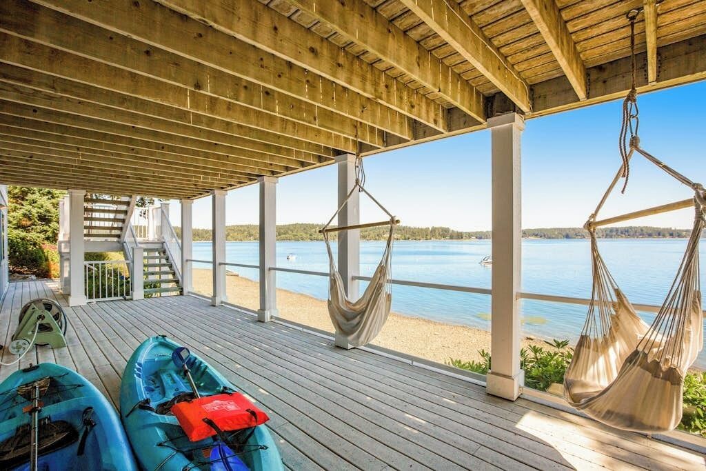 A covered deck with a view of a lake, two kayaks, and a hammock.