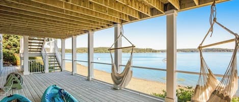A covered deck with a view of a lake, two kayaks, and a hammock.