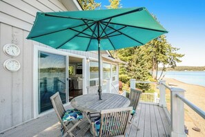 Una terraza de madera con una mesa, sillas, un paraguas verde y un ventanal que da a un espacio interior.