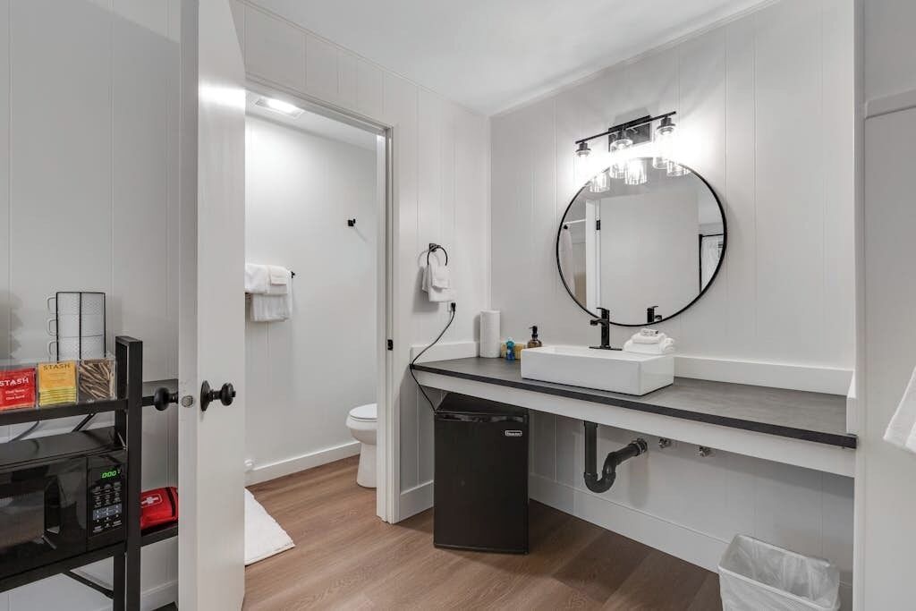 A modern bathroom with a white sink, a round mirror, and a black trash bin.