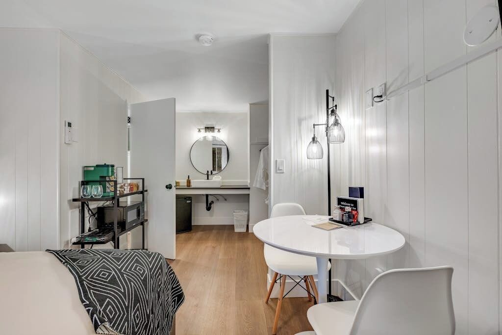 A compact hotel room with a round dining table, a white sofa, a small kitchenette, and a mirror with lights above the sink.