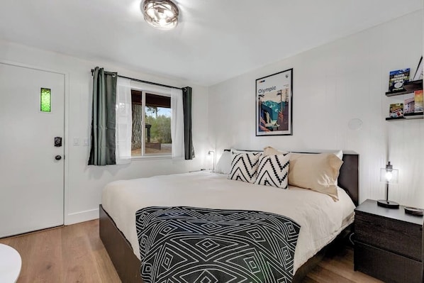 Een slaapkamer met een bed, een nachtkastje, een raam met gordijnen, een deur en een aan de muur bevestigd schilderij.