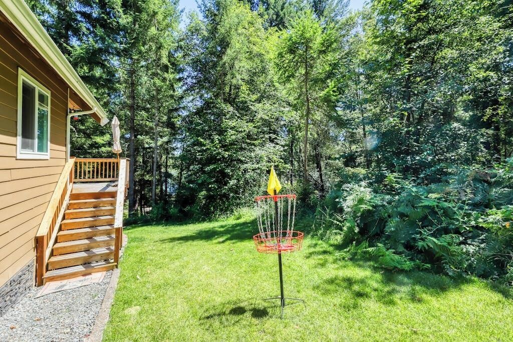 Casa de madera con porche delantero y un cesto de disc golf en un patio cubierto de césped, rodeado de árboles.