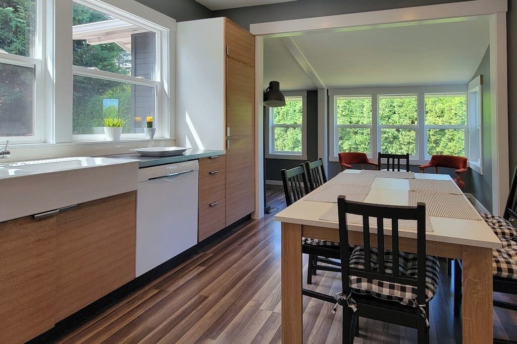 A modern kitchen with wooden cabinets, a dining area with a long table and chairs, and large windows offering a view of greenery.