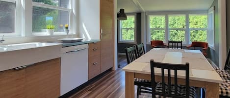 A modern kitchen with wooden cabinets, a dining area with a long table and chairs, and large windows offering a view of greenery.