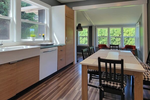 Een moderne keuken met houten kastjes, een eethoek met een lange tafel en stoelen, en grote ramen met uitzicht op groen.