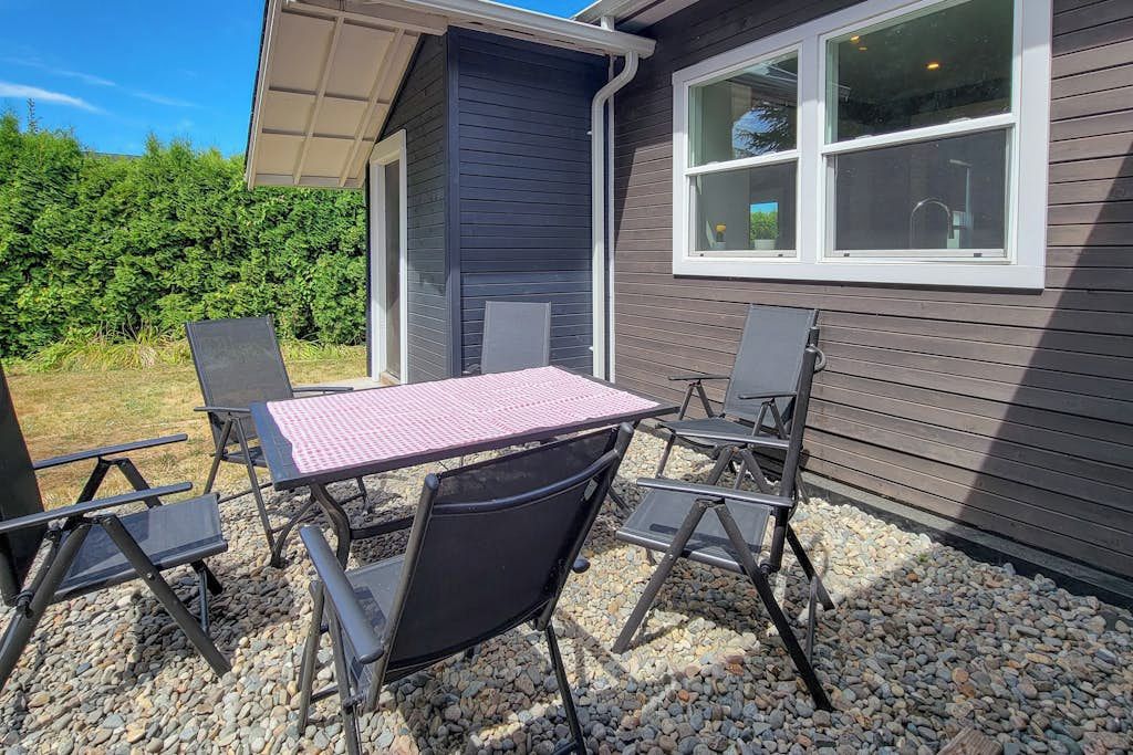 An outdoor seating area with a table and chairs, a window, and a hedge in the background.