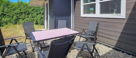 An outdoor seating area with a table and chairs, a window, and a hedge in the background.