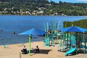 A beach with a playground, including slides and climbing structures, and a view of a lake with houses in the background. - 2BR charmer near the lake with large yard & bike (Renton)