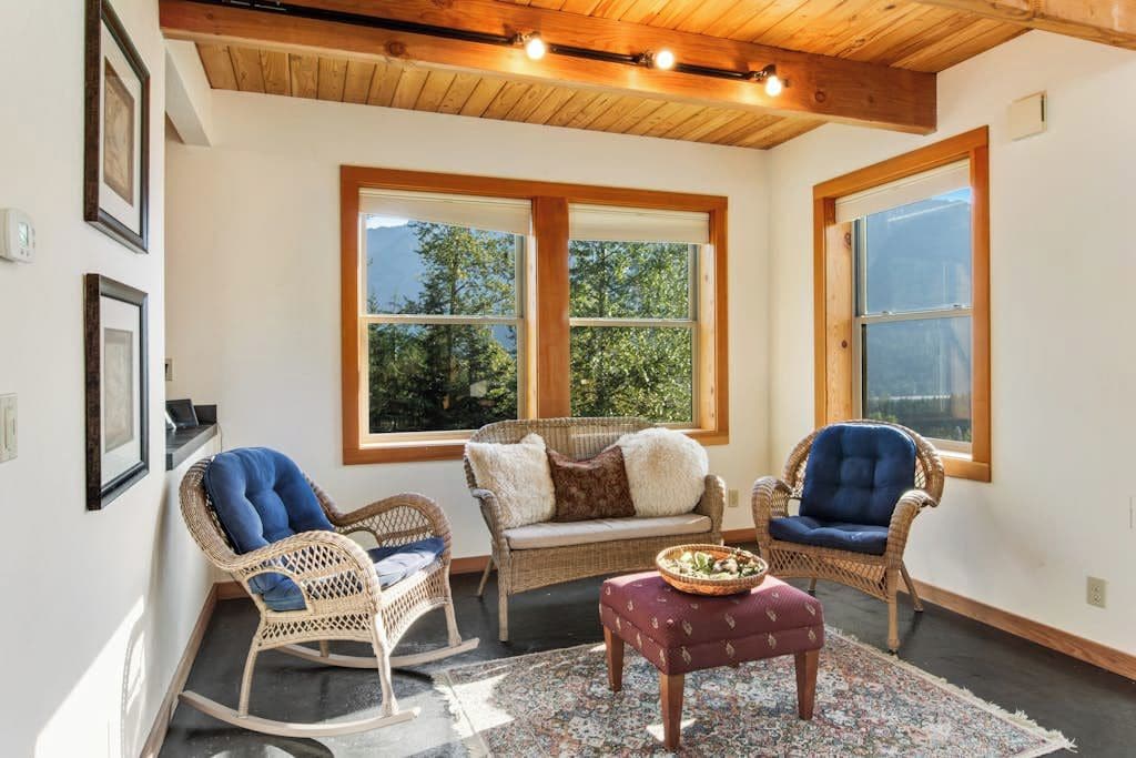 A room with wicker furniture, a patterned rug, and large windows with a view of trees.