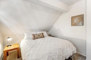 Een slaapkamer met een bed, nachtkastje, lamp en een aan de muur hangend schilderij.