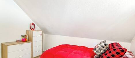 A bedroom with a sloped ceiling, a bed with red bedding, a wooden dresser, and a red lantern.