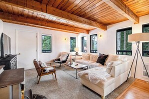 A modern living room with a wooden ceiling, a white sofa, a coffee table, and a TV. - 4BR - Sauna - WoodStove - Balcony - Firepit - Washer Dryer (Snoqualmie Pass)