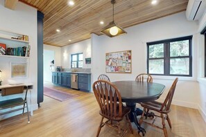 A dining area with a round table and wooden chairs, a kitchen in the background, and a bulletin board on the wall. - Cabin 3BR - Foosball - Gas Grill - Kids Games (Ronald)