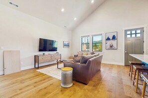 A spacious living room with a flat-screen TV, a brown leather sofa, a dining area with a table and chairs, and large windows offering a view of the outdoors. - 3 BR Home with loft, game room, fire table & trails (Cle Elum)