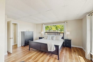 A bedroom with a bed, a dresser, a nightstand, a window with a view of greenery, and a door. - 4BR Bayview - Gas Fire Pit - Balcony - Washer Dryer (Bainbridge Island)