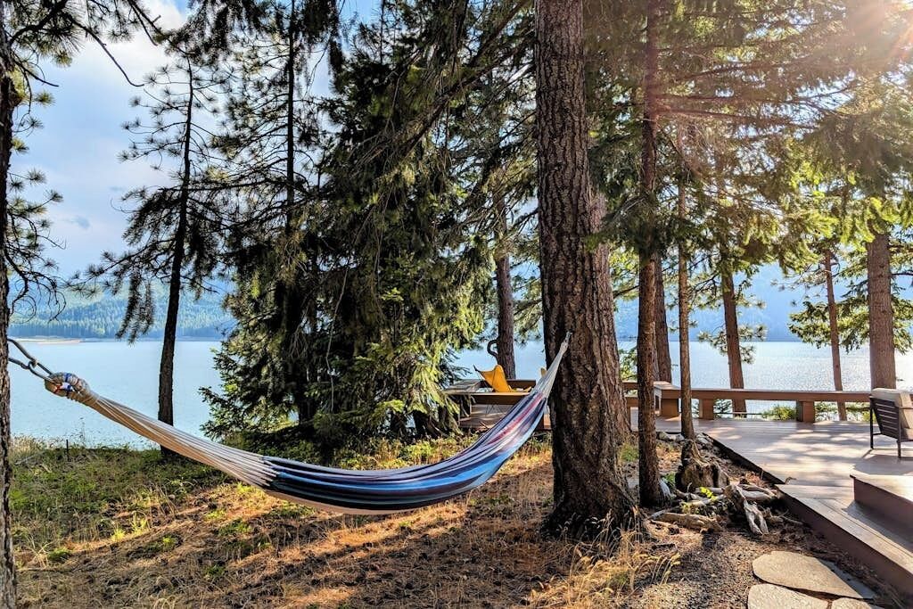 A hammock tied between trees overlooking a lake with a wooden deck and chairs.