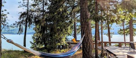 A hammock tied between trees overlooking a lake with a wooden deck and chairs.
