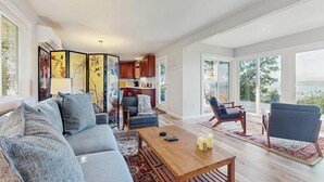 Amplia sala de estar con sofá, mesa de centro y sillas, una cocina al fondo y grandes ventanales con vistas.