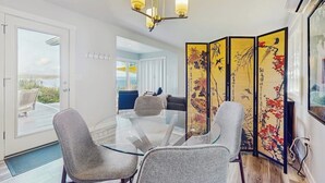 Un comedor moderno con una mesa de vidrio, sillas tapizadas en gris, una lámpara decorativa en el techo y un biombo de estilo asiático con obras de arte.