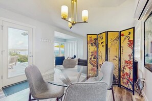 Un comedor moderno con una mesa de vidrio, sillas tapizadas en gris, una lámpara decorativa en el techo y un biombo de estilo asiático con obras de arte.