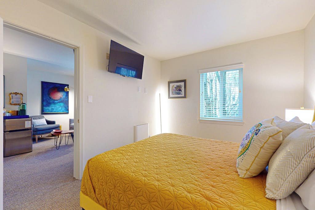 A bedroom with a large bed, a television mounted on the wall, a window with blinds, and a door leading to a living area.