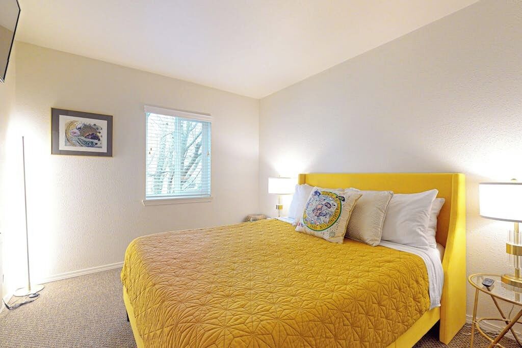 A hotel room with a bed, a bedside table, a lamp, a window, and a framed picture on the wall.