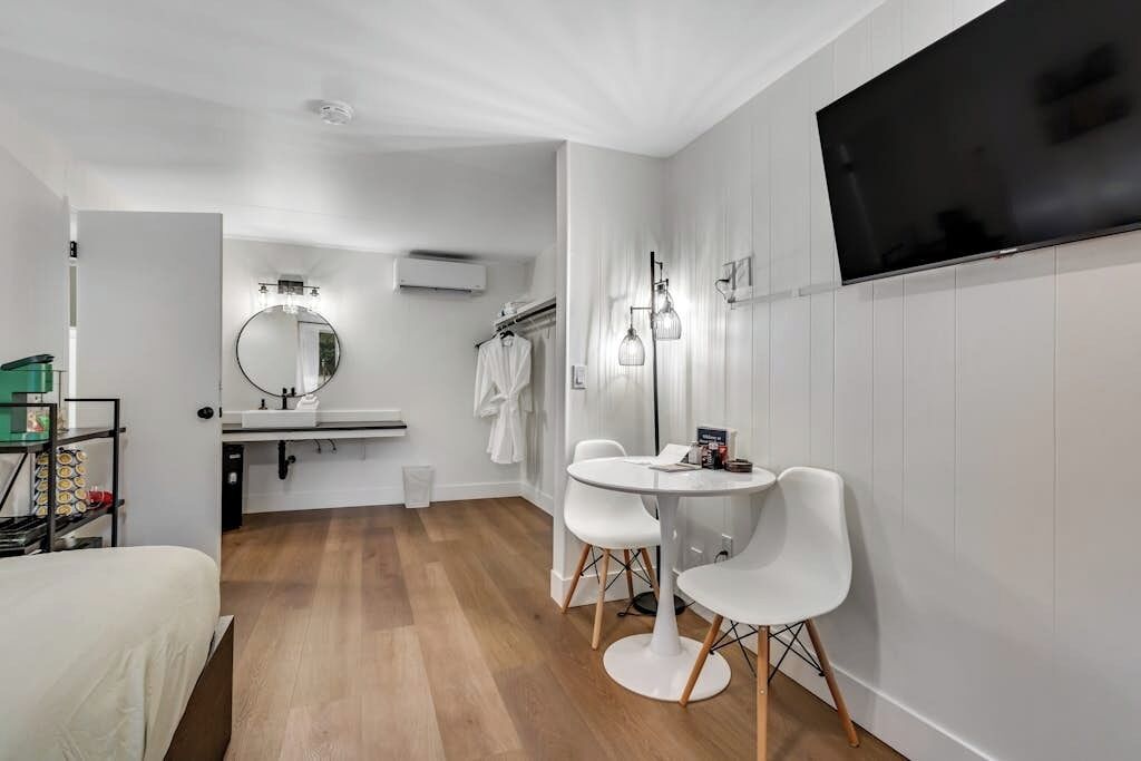 Una habitación de hotel moderna con una cama, un espejo redondo, un lavabo blanco, una mesa redonda con dos sillas y un televisor de pantalla plana montado en la pared.