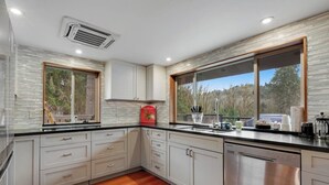 A modern kitchen with white cabinets, a stainless steel dishwasher, and a large window offering a view of trees.