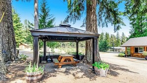 Un kiosque en bois avec une table de pique-nique, entouré de grands arbres et avec un bâtiment visible au loin.