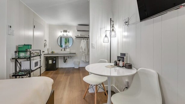 A modern hotel room with a round dining table, white chairs, a small kitchenette, a round mirror, and a flat-screen TV mounted on the wall.