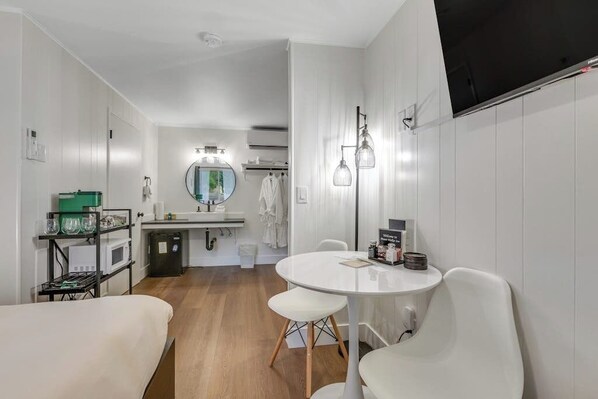 Quarto de hotel moderno com uma mesa de jantar redonda, cadeiras brancas, uma pequena cozinha compacta, um espelho redondo e uma TV de tela plana fixada na parede.