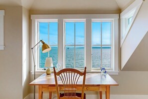 Quarto com uma mesa de madeira, uma cadeira, um abajur e vista para o mar através de grandes janelas.