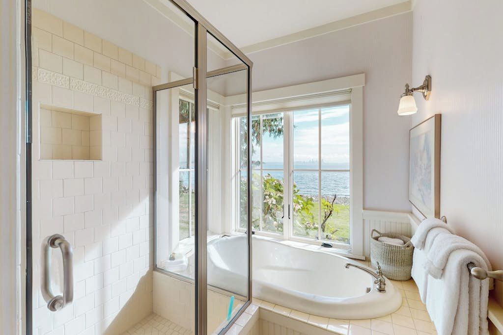 A bathroom with a glass-enclosed shower, a bathtub, and a large window offering a view of the outdoors.