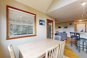 Ein Essbereich mit Tisch und Stühlen, ein Fenster mit Jalousien, eine Couch und eine Küchenzeile im Hintergrund.