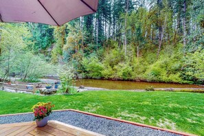 A patio with a view of a forested area and a stream. - 2BR home with hot tub, firepit, Washer Dryer, riverfront (Tahuya)