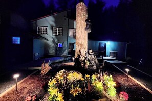A nighttime view of a house with a garden featuring a large tree stump and flowering plants. - 2BR home with hot tub, firepit, Washer Dryer, riverfront (Tahuya)
