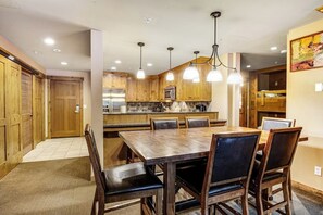 A dining area with a wooden table and chairs, a kitchen with wooden cabinets and a microwave, and pendant lights above the table.