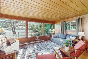 A cozy room with a wooden ceiling, large windows, a sofa, a chair, and a small table with a plant. - 1BR Soundview House - Fireplace (Gig Harbor)