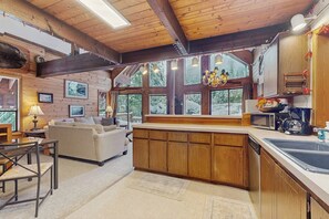 Een interieur van een blokhut met houten plafond, een eethoek, een bank, een keuken met spoelbak en grote ramen.