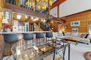 A spacious living area with a high ceiling, wooden beams, a glass dining table, and a sofa. - Delightful 2BR - WoodStove - Balcony - Game Room (Snoqualmie Pass)