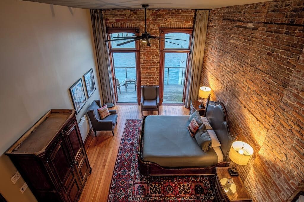 Een kamer met een bed, een bureau, een stoel en uitzicht op zee door grote ramen.