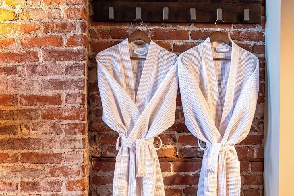 Two white bathrobes hanging on a rack against a brick wall.