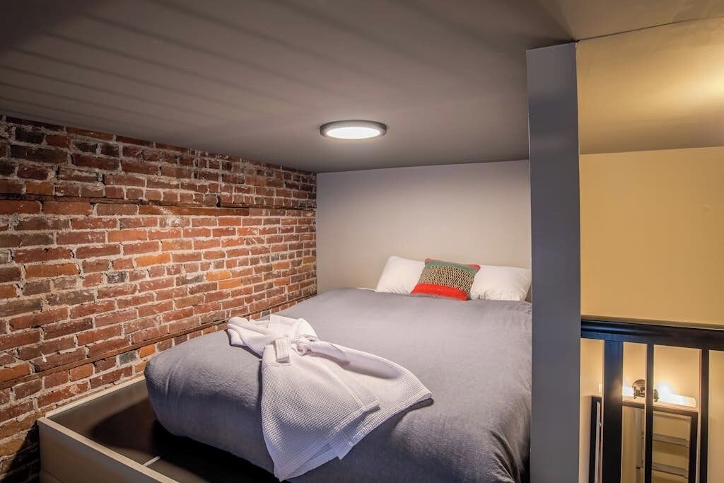 A hotel room with a brick wall, a bed with a grey bedspread, a white robe, and a small bedside table with a lamp.