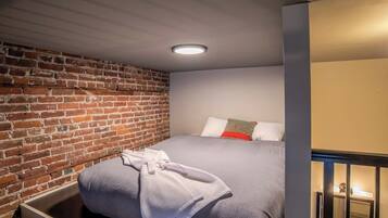 A hotel room with a brick wall, a bed with a grey bedspread, a white robe, and a small bedside table with a lamp.