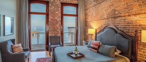 A hotel room with a large bed, two chairs, a wooden floor, a brick wall, and a balcony with a view.