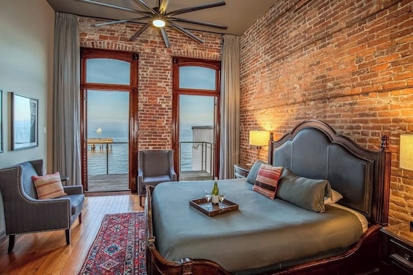 A hotel room with a large bed, two chairs, a wooden floor, a brick wall, and a balcony with a view. - Studio Room w/ Water Views overlooking The Bay (Port Townsend)